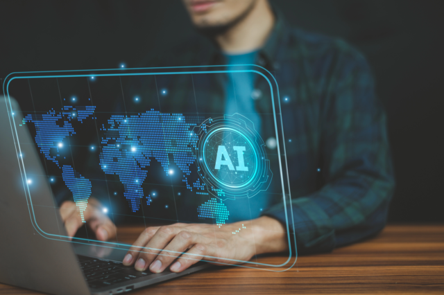 Holographic display showing a digital world map with glowing AI icon overlay and person typing on laptop